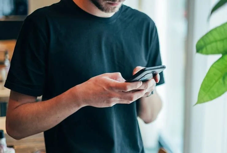A person wearing a black t-shirt is standing indoors, holding and using a smartphone near a window.