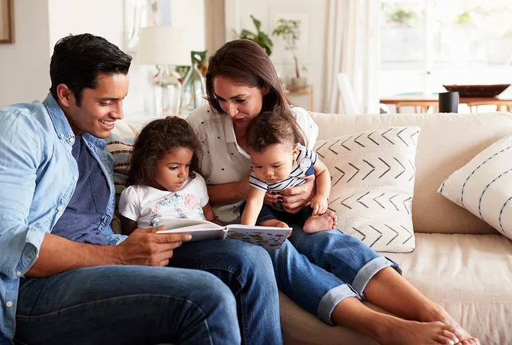 A family of four sitting together on a couch, reading a book