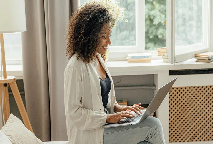 A woman sitting on a bed, focused on her laptop in a relaxed bedroom environment